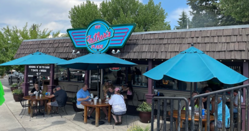 A restaurant with a retro sign, outdoor seating under blue umbrellas, and diners enjoying meals on a sunny day.