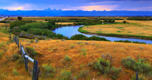 A serene landscape featuring a winding river, grassy fields, and distant mountains under a colorful sky.
