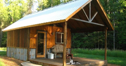 A rustic wooden cabin with a metal roof, surrounded by trees, featuring a porch and a dog resting on the ground.