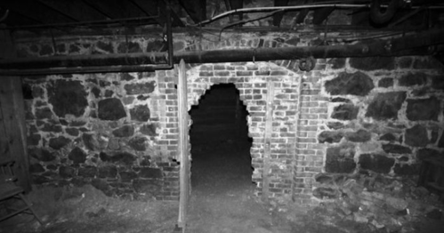 A dimly lit stone basement with a partially collapsed brick archway leading into darkness.