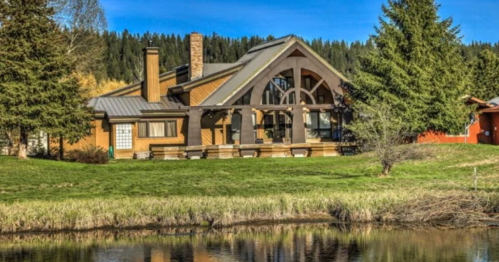A large, modern cabin with a sloped roof, surrounded by trees and reflecting in a calm lake under a clear blue sky.