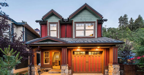 A charming two-story house with a red exterior, green accents, and a stone porch, surrounded by trees.