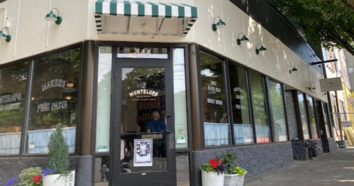 Exterior view of Montelupo, a restaurant with large windows and a striped awning, surrounded by greenery and flowers.