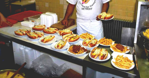 A kitchen counter with multiple plates of hot dogs and fries, with a person holding two plates in the background.