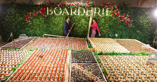 A vibrant display of assorted desserts on large wooden boards, with a floral backdrop and two people interacting.