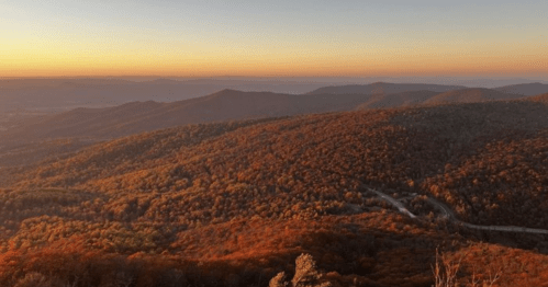 A scenic view of rolling mountains covered in autumn foliage at sunset, with warm colors illuminating the landscape.