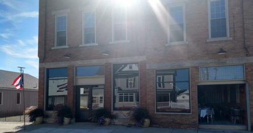 A sunlit brick building with large windows and potted plants, featuring a flag in the background.