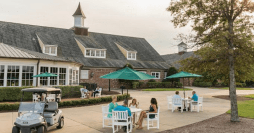 A golf club patio with people dining at tables under green umbrellas, and a golf cart parked nearby.