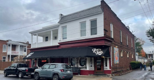 A two-story brick building with a black awning reading "Joe's Jan," surrounded by parked cars and cloudy skies.
