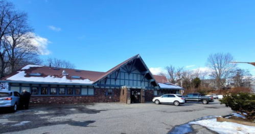 A large, rustic building with a sloped roof, surrounded by a parking lot and trees, under a clear blue sky.