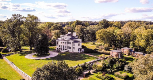 Aerial view of a large white mansion surrounded by lush greenery and gardens under a blue sky with fluffy clouds.