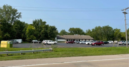 A parking lot filled with vehicles next to a single-story building, surrounded by trees under a clear blue sky.