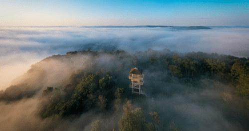 Aerial view of a solitary tower surrounded by dense fog and lush greenery at sunrise.