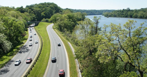 A winding road alongside a river, surrounded by lush greenery and trees under a clear blue sky.