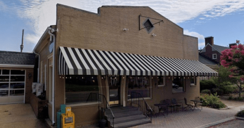 A brick building with a striped awning, outdoor seating, and a newspaper stand in front, under a blue sky.