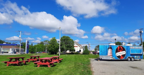 A food truck serving fresh seafood parked near red picnic tables under a blue sky with fluffy clouds.