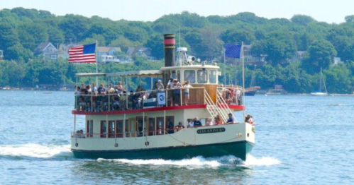 A vintage-style boat with passengers cruising on a calm waterway, surrounded by green trees and distant sailboats.