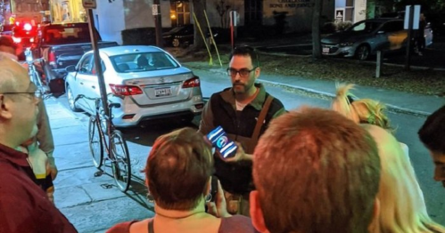 A group of people gathered on a street, watching a man holding a phone, illuminated by nearby lights.