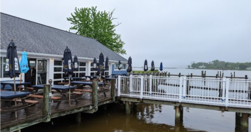 A waterfront restaurant with outdoor seating and umbrellas, overlooking a calm body of water on a cloudy day.