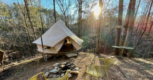 A cozy tent in a wooded area, with a fire pit and yellow chairs, surrounded by trees and sunlight filtering through.