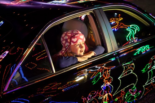 A child with a heart-patterned hat gazes out of a car window, surrounded by colorful holiday lights.