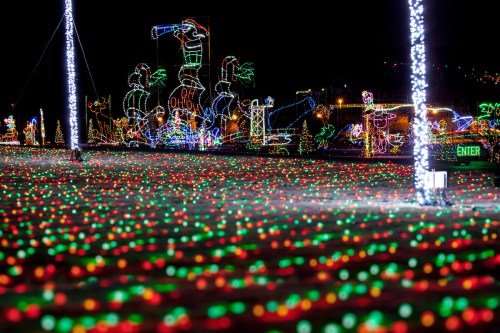 A colorful display of holiday lights covers the ground, with festive decorations and illuminated figures in the background.