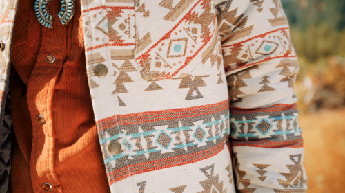 Close-up of a patterned jacket with geometric designs, worn over an orange shirt, set against a natural background.
