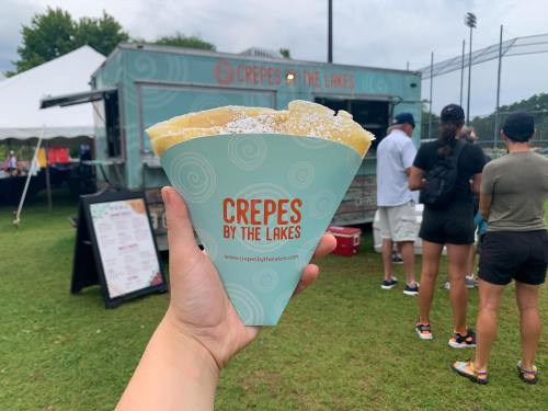 A hand holds a cone of crepes in front of a food truck labeled "Crepes by the Lakes," with people in the background.