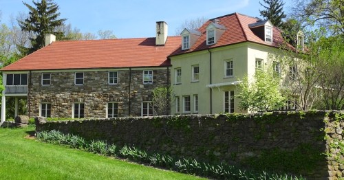 A large, two-story house with a mix of stone and yellow siding, surrounded by greenery and a stone wall.