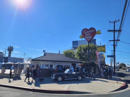 A busy hot dog stand with a vintage car parked outside, people waiting in line under a clear blue sky.