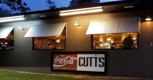 A dimly lit building with open windows, featuring a Coca-Cola sign and a sign that reads "CUTTS."