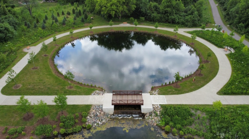 Aerial view of a circular pond surrounded by pathways and greenery, featuring a small bridge over a stream.