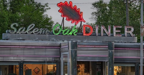 Neon sign of Salem Oak Diner with a leaf design, set against a backdrop of trees and a cloudy sky.