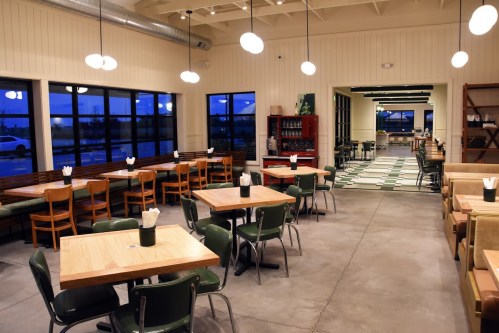 A modern restaurant interior with wooden tables, green chairs, and large windows, illuminated in the evening.