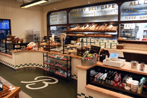 A cozy bakery deli cafe featuring a variety of breads, pastries, and beverages displayed on wooden shelves and counters.