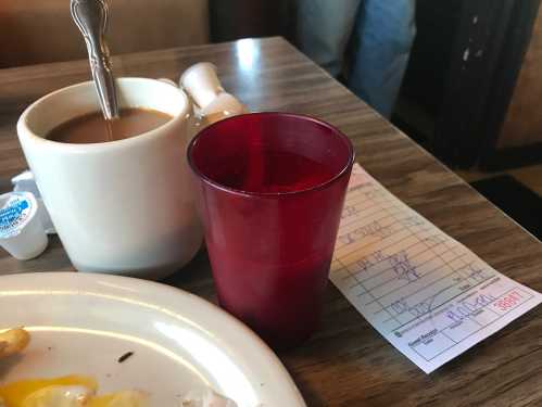 A cup of coffee, a red plastic glass, and a receipt on a table with a plate of food in a diner setting.