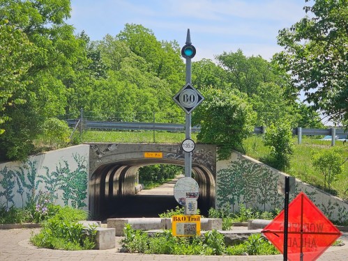 A green-lit tunnel entrance with B&O Trail signs, surrounded by lush greenery and a warning sign in the foreground.