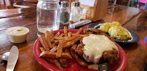 A plate of fried chicken topped with cheese, served with fries and a side of scrambled eggs, alongside a glass of water.