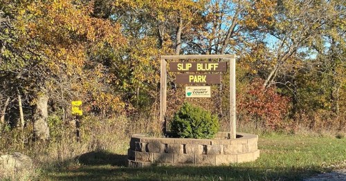 Sign for Slip Bluff Park surrounded by trees with autumn foliage. Clear blue sky in the background.