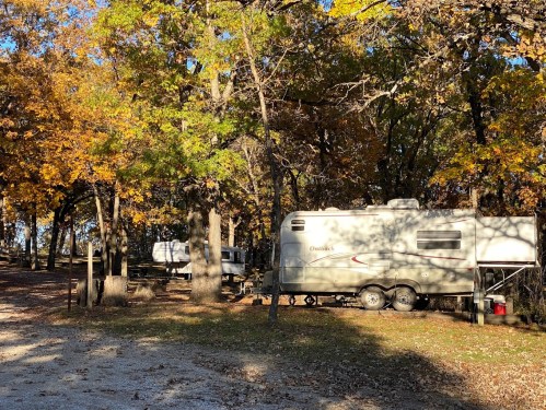 A camper parked among colorful autumn trees, with a gravel path and another trailer in the background.