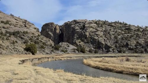 A winding river flows through a grassy landscape, flanked by rugged rock formations and sparse trees under a blue sky.