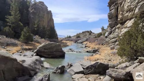 A serene river flows between rocky cliffs, surrounded by trees and dry grass under a clear blue sky.