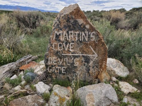 A weathered rock sign pointing to "Martin's Cove" and "Devil's Gate," surrounded by shrubs and rocky terrain.