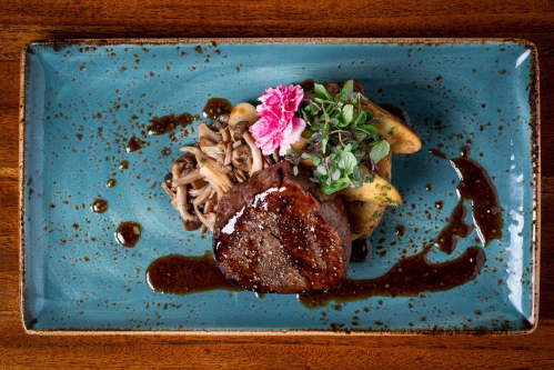 A beautifully plated dish featuring a steak, mushrooms, and garnishes on a blue plate with a drizzle of sauce.