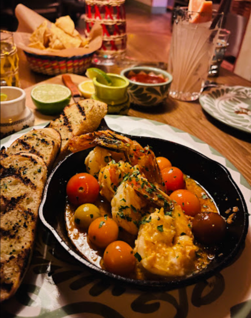 A skillet dish with shrimp, colorful tomatoes, and herbs, served with grilled bread and sides of salsa and lime.