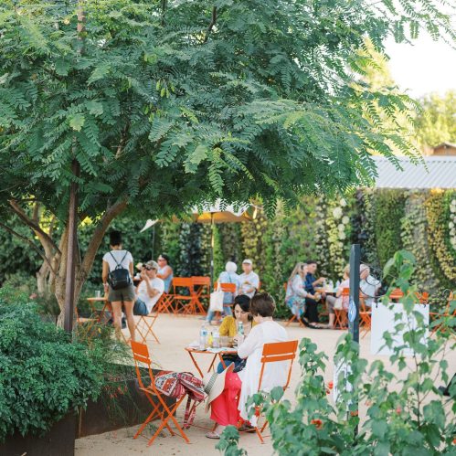 A vibrant outdoor café scene with people sitting at orange chairs under lush greenery and floral decorations.