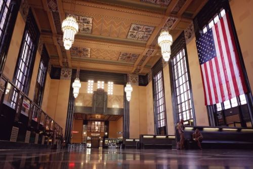 Interior of a grand hall with Art Deco design, featuring chandeliers, large windows, and an American flag.