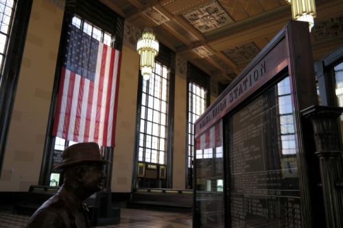 Interior of a train station featuring a large American flag, vintage decor, and a schedule board with a statue in the foreground.