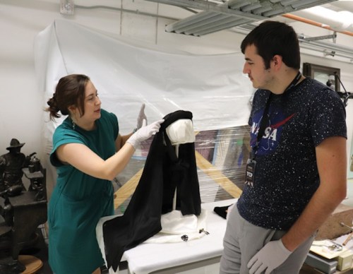 A woman in a green dress demonstrates a garment to a man in a NASA shirt in a workshop setting.