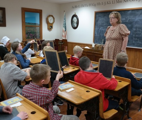 A teacher instructs a classroom of children holding chalkboards in a vintage school setting.